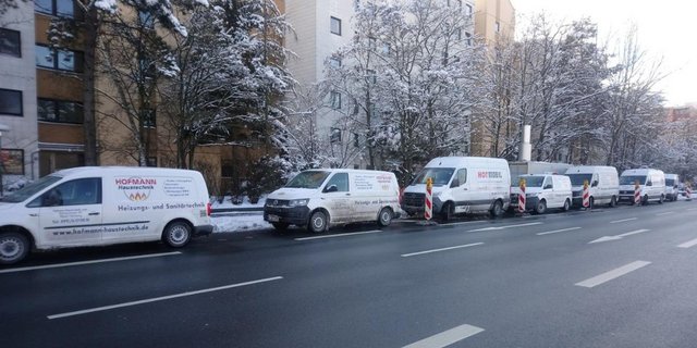Eine Reihe von Handwerkerfahrzeugen parkt auf einer schneebedeckten Straße, umgeben von winterlichen Bäumen und Gebäuden.