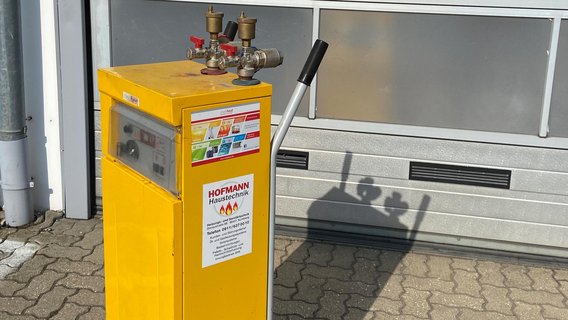 Eine gelbe Gasflasche mit Ventilen steht vor einer grauen Wand, während die Sonne einen klaren Schatten wirft.