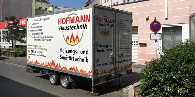 Ein Anhänger mit Werbung für Heizungs- und Sanitärtechnik steht an einer Straße, umgeben von Wohngebäuden und Bäumen.
