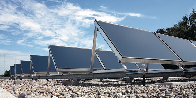 Eine Reihe von Solarpanelen steht auf einem Dach, umgeben von Kies, unter einem klaren blauen Himmel mit wenigen Wolken.