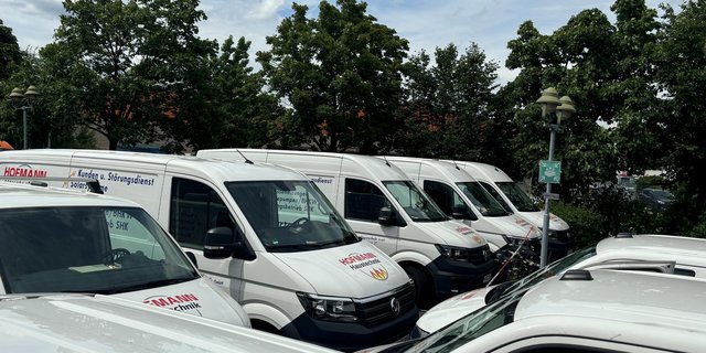 Eine Reihe von weißen Transportern ist in einer Parklücke angeordnet, umgeben von grünen Bäumen und einem Notausgangsschild.