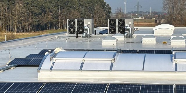 Auf dem Dach sind mehrere Solarpanels und Lüftungsanlagen zu sehen, umgeben von einer ländlichen Landschaft.