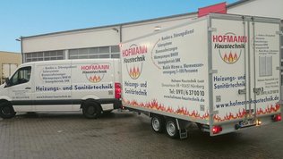 Ein weißer Transporter und ein Anhänger mit dem Logo von Hofmann Haustechnik stehen auf einem gepflasterten Parkplatz.
