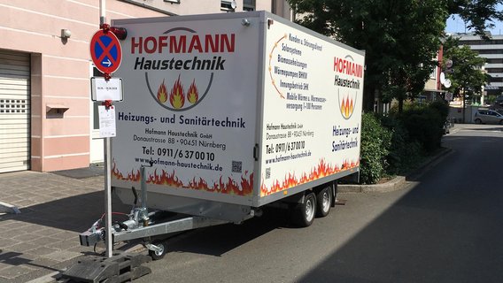 Ein Anhänger mit dem Logo von Hofmann Haustechnik steht an einer Straße, umgeben von Bäumen und Gebäuden.
